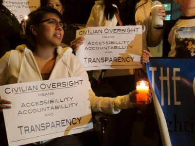 Los Angeles native, Aloni Bonillas holds a candle and sign beside several other community members in front of the County jail, during Wednesday night’s vigil. (Neon Tommy / Celeste Alvarez)